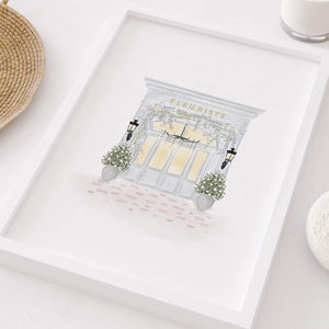 Chic Parisian 'Fleuriste' flower shop watercolor print in a white frame, placed beside a basket and a modern vase, enhancing a stylishly minimalistic home decor, by artist Leah Straatsma