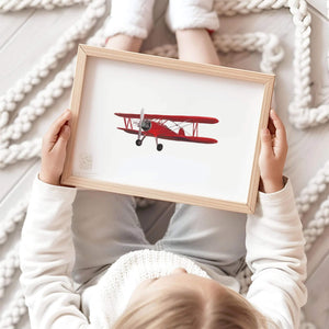 Child in a cozy sweater holding a framed art print of a classic red biplane, adding a touch of vintage aviation charm to a serene nursery setting, by artist Leah Straatsma